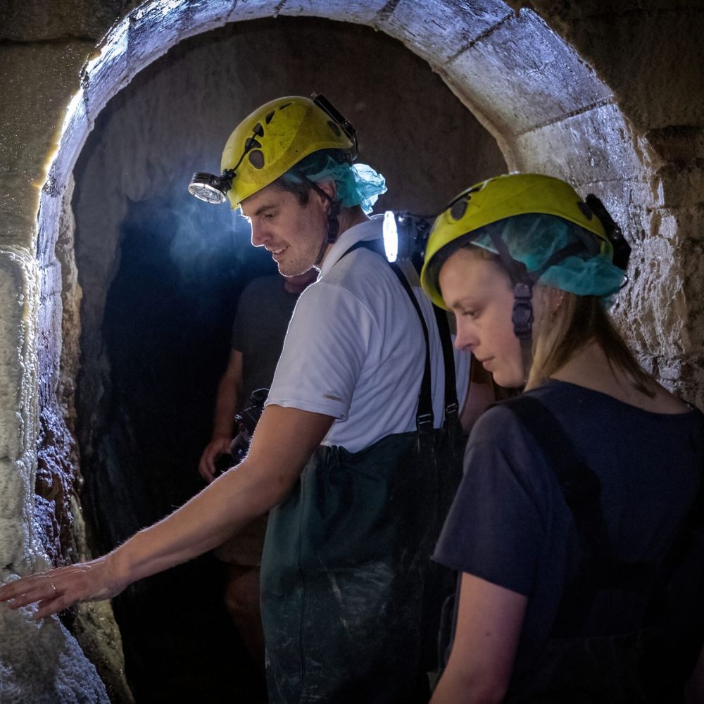 Two people explore a dimly lit cave, wearing yellow helmets with headlamps and protective clothing. One person touches the stone wall, while the other observes, both focused on their surroundings.