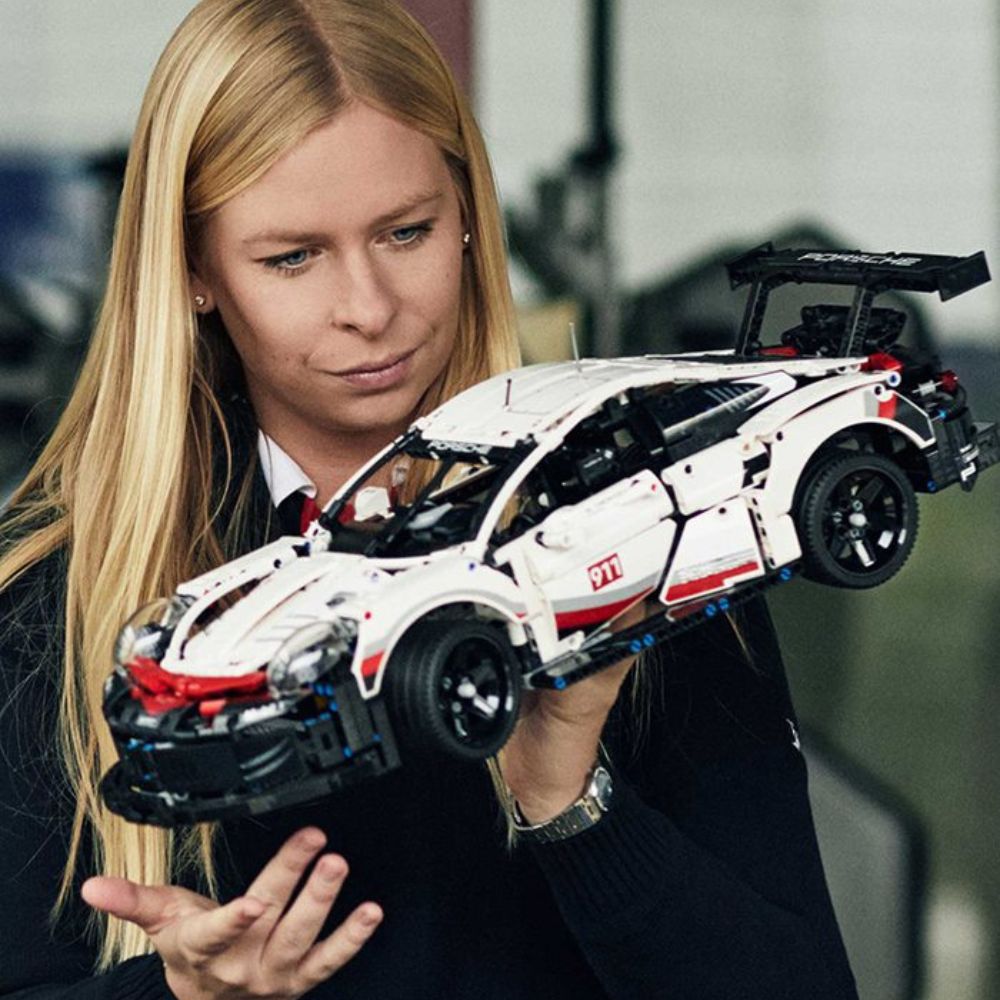 A woman with long blonde hair examines a detailed model of a white sports car made from building blocks. She wears a black blazer and appears focused on the model, which features intricate design elements and a prominent rear wing.