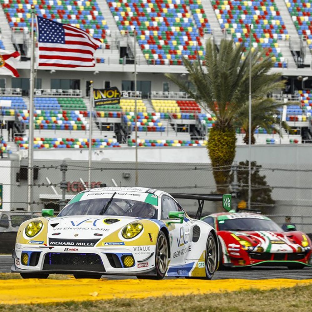 A yellow and white race car with sponsor logos speeds along a racetrack, while a red race car follows closely behind. Colorful grandstands filled with spectators and flags in the background enhance the vibrant atmosphere of the racing event.