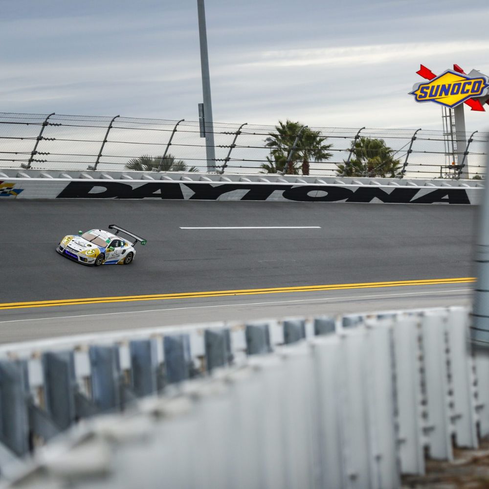 A sports car speeds around the Daytona International Speedway, with palm trees and a clouded sky in the background. The Sunoco logo is visible in the corner.