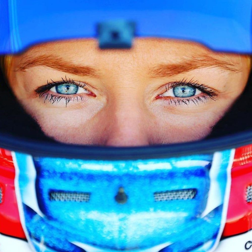 Close-up of a race car driver's face, featuring striking blue eyes peering from inside a helmet. The helmet displays a vibrant design with elements of blue and red.
