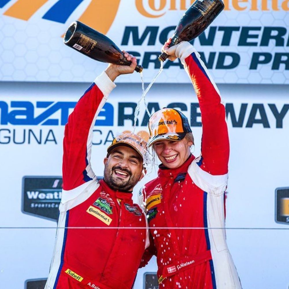 Two jubilant race car drivers celebrating on the podium, spraying champagne in the air after a victory at the Monterey Grand Prix. They are wearing red racing suits and baseball caps with sponsor logos.