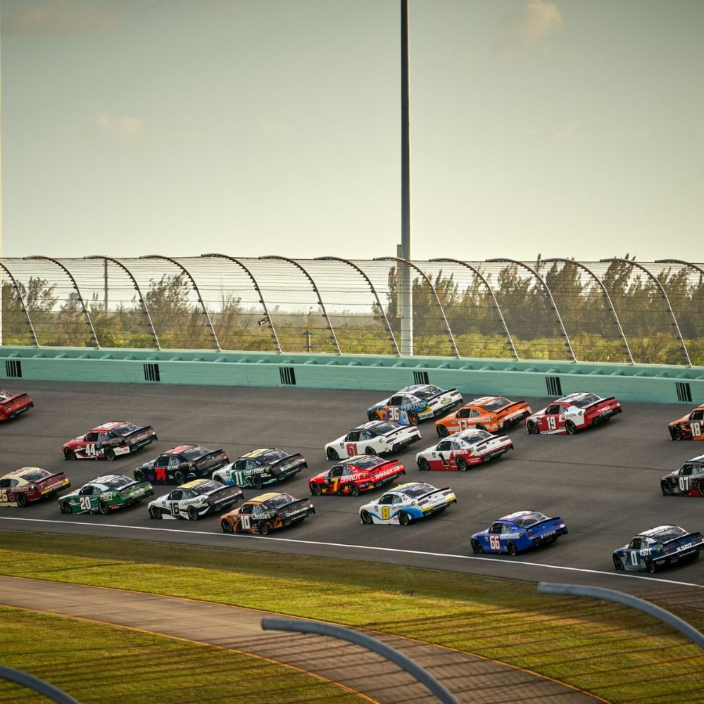 Race cars speed around a banked track during a NASCAR event at a stadium. The scene captures a variety of colorful vehicles and a clear blue sky in the background, showcasing the intensity of the competition.