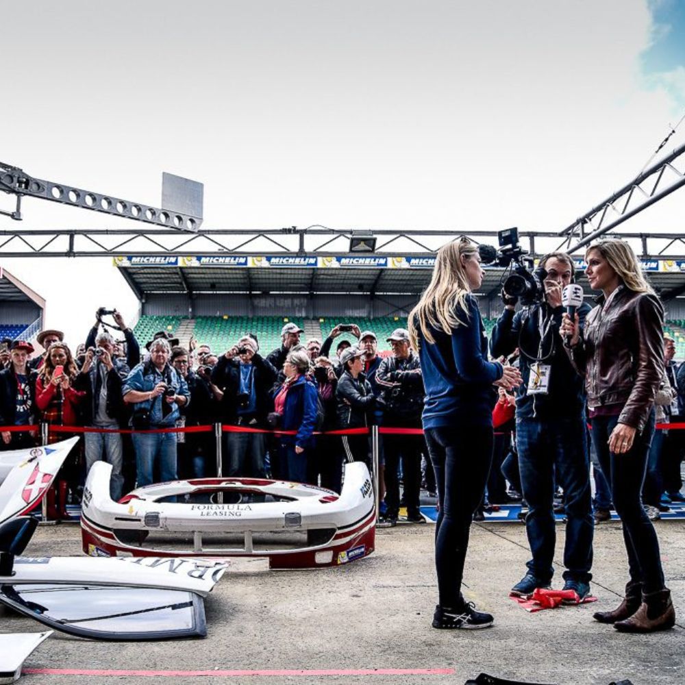 A motorsport event scene with a large crowd of spectators. Two women are being interviewed, one holding a microphone and wearing a leather jacket. A race car is visible in the foreground, with its parts scattered.