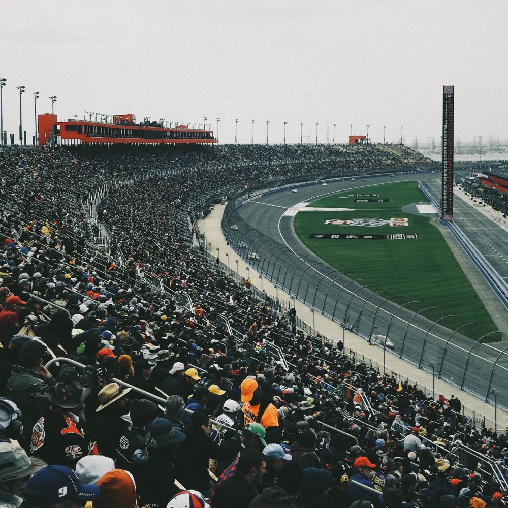 A large crowd fills the grandstands at a racetrack, overlooking the track and pit area. The stands are packed with fans, many wearing caps and shirts in support of various racing teams. The atmosphere is vibrant and energetic, with a mix of cloudy skies above.