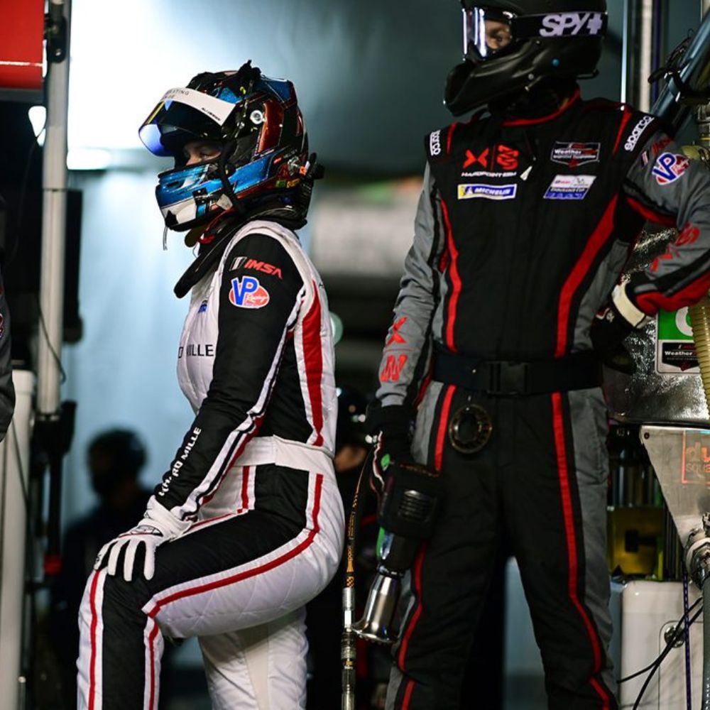A female race car driver in a white racing suit sits on a pit wall, wearing a helmet and looking focused. Beside her, a male team member in a black jumpsuit stands with a helmet, both awaiting a race.