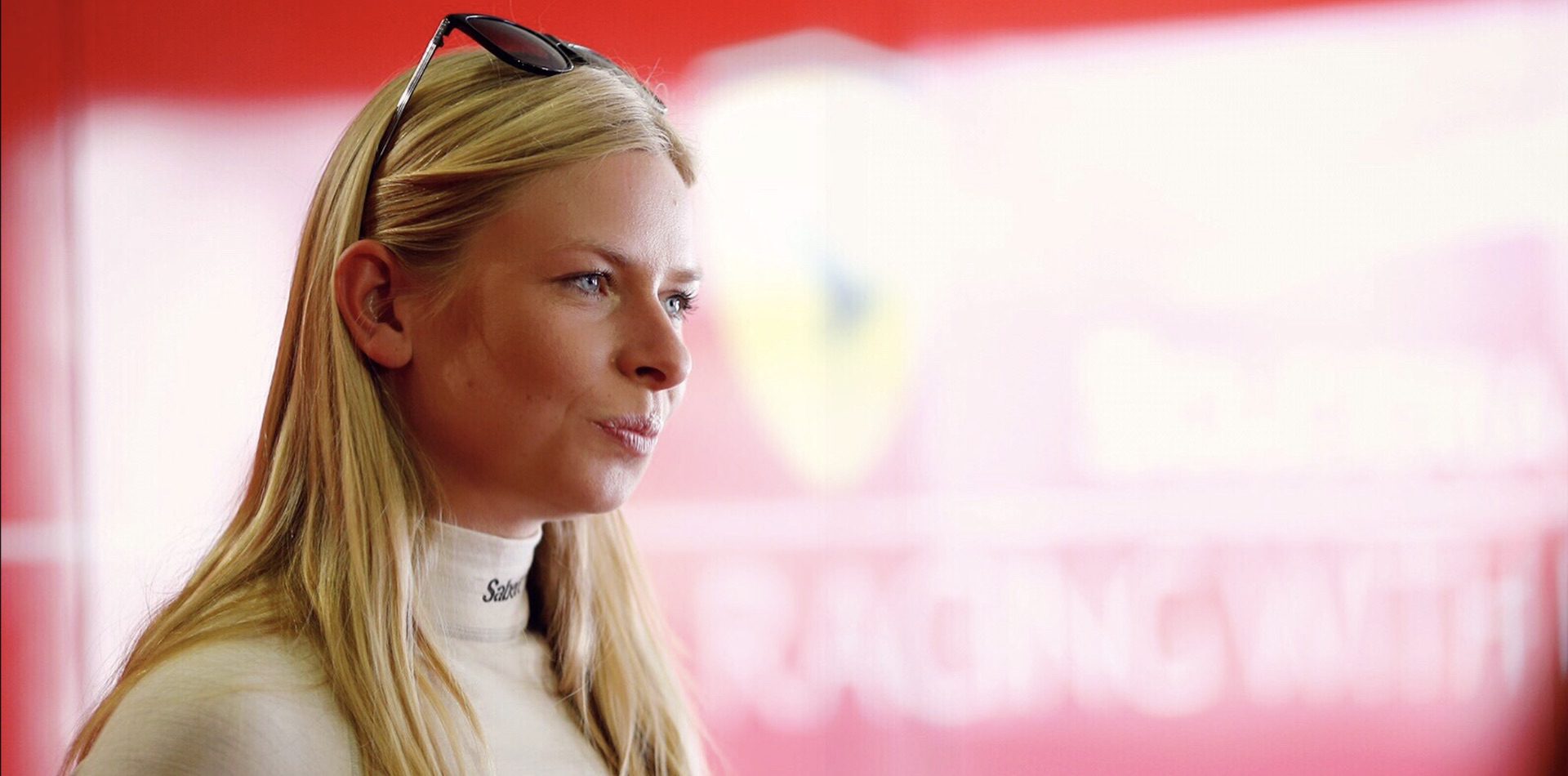 A young woman with long blonde hair is wearing a racing suit, standing in front of a red backdrop with a Ferrari logo. She has sunglasses perched on her head and is looking off to the side, with a thoughtful expression.