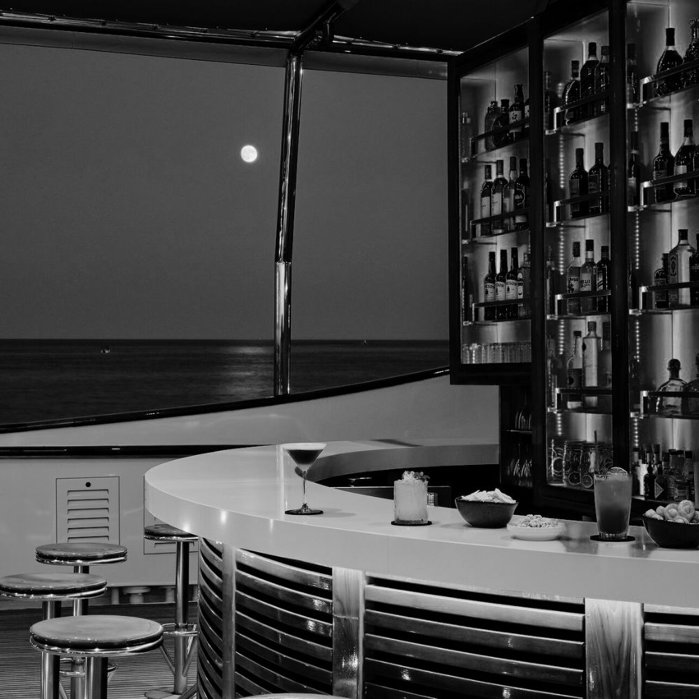 Black and white photo of a sleek outdoor bar on a yacht at night with cocktails on the counter and the moon reflecting on the sea.