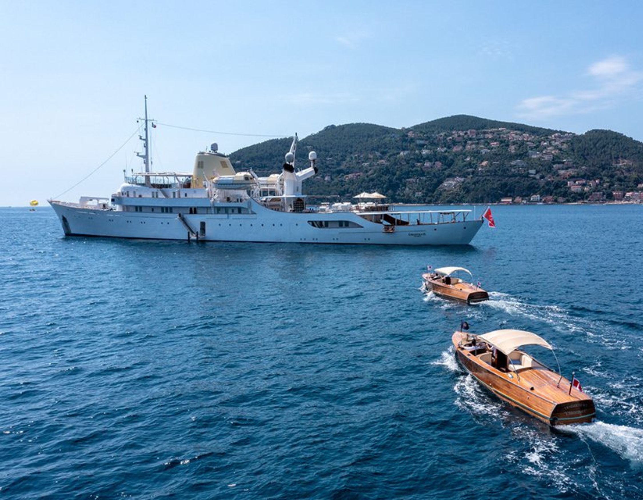 A large white yacht anchored in calm blue waters, with two wooden boats approaching it. A lush green hillside with buildings is visible in the background under a clear blue sky.