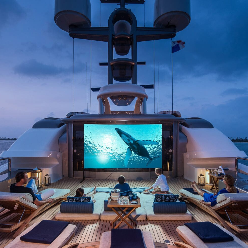 People lounge on the deck of a yacht at twilight, watching a movie of a whale on a large outdoor screen.