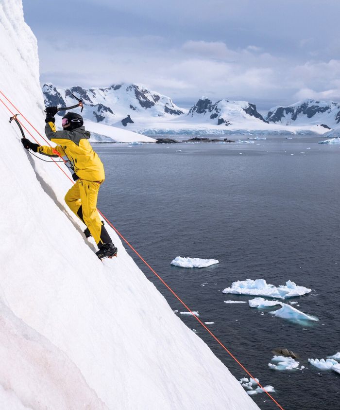 An ice climber in a yellow suit scales a vertical white ice wall overlooking a cold sea with icebergs and snowy mountains.