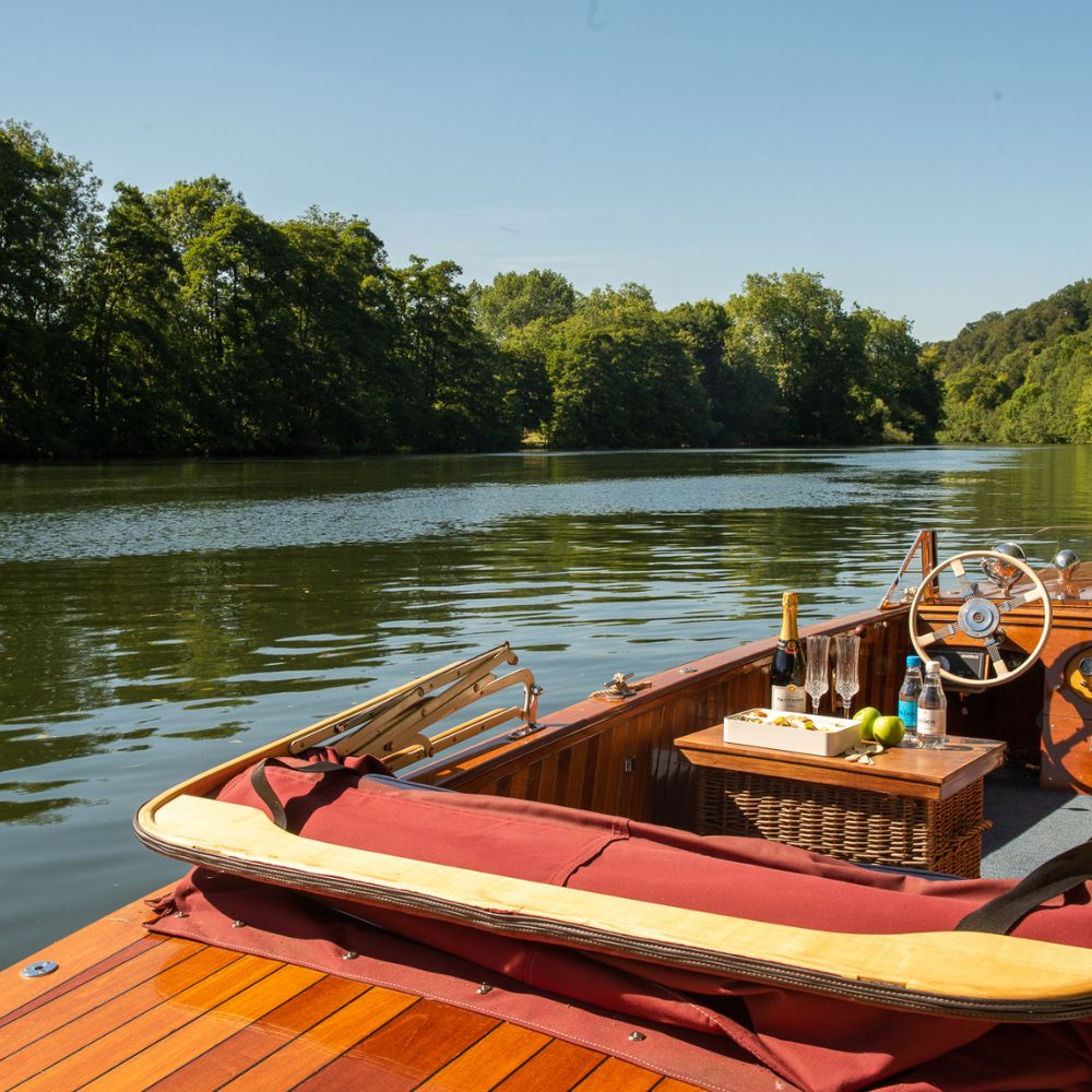 A wooden boat on calm water, surrounded by lush greenery. The boat features a wicker basket and a bottle of champagne on a table, with apples nearby. Bright blue sky visible above.