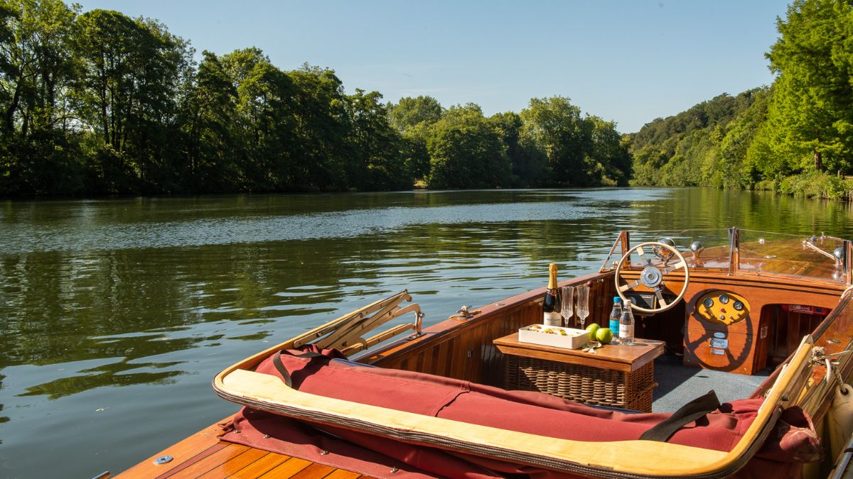 A vintage wooden boat floats on a serene river, surrounded by lush green trees and a clear blue sky. A small table is set up on the boat with a picnic basket, drinks, and limes. The calm water reflects the greenery, creating a peaceful atmosphere.