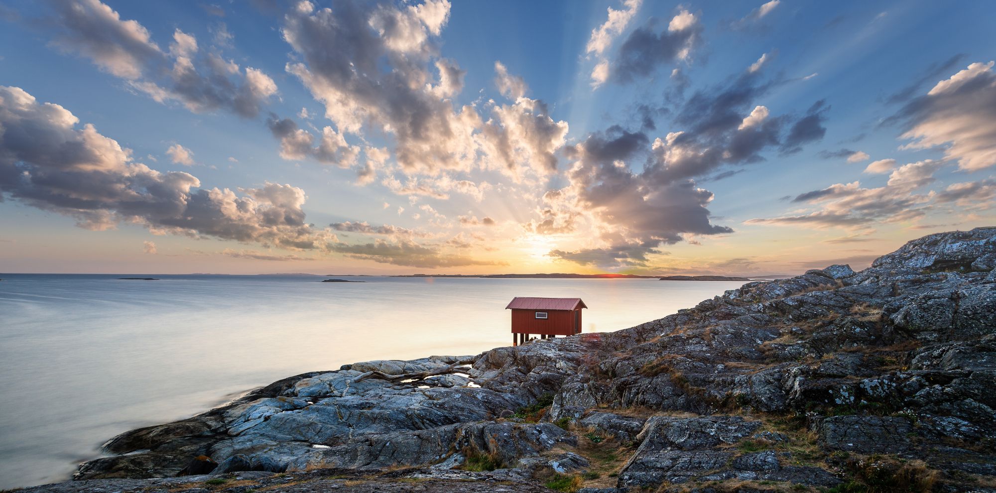 A small red cabin on rocky shoreline at sunset, with dramatic clouds and soft light reflecting on calm water.