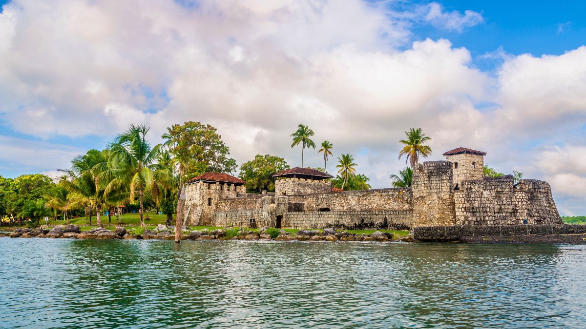 Historic stone fortress surrounded by palm trees and water, with a cloudy blue sky in the background.
