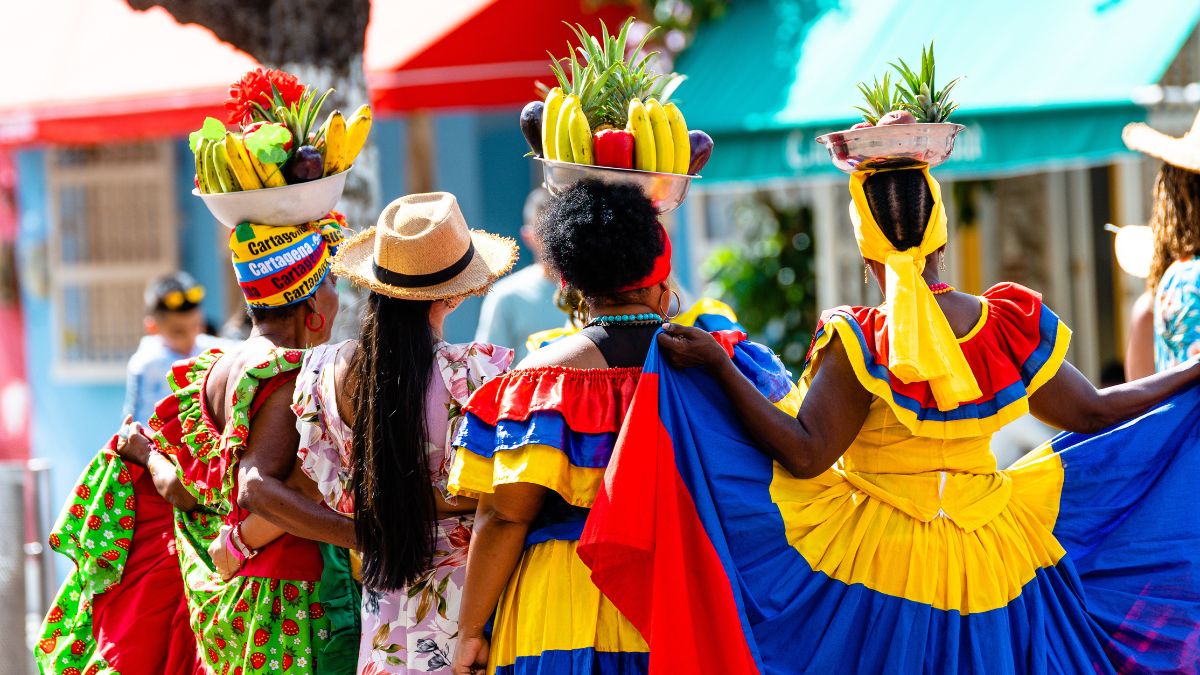 Colorful festival-goers wear vibrant traditional dresses and large hats, carrying baskets of fruits on their heads while joyfully walking together in a lively outdoor setting.