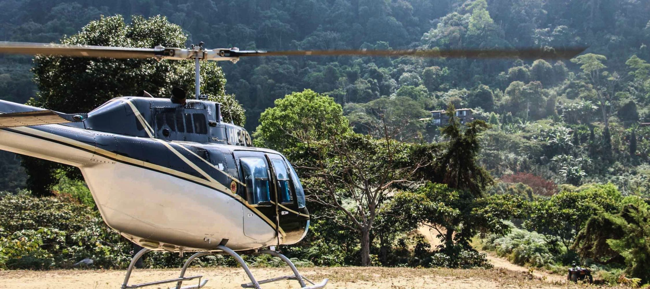 A helicopter is parked on a dirt landing area surrounded by lush green trees and hills. The landscape features dense foliage in the background.