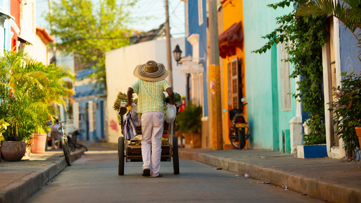 A person wearing a straw hat walks down a colorful street, pushing a cart filled with goods. Brightly painted buildings line the narrow street, with greenery and bicycles visible in the background.