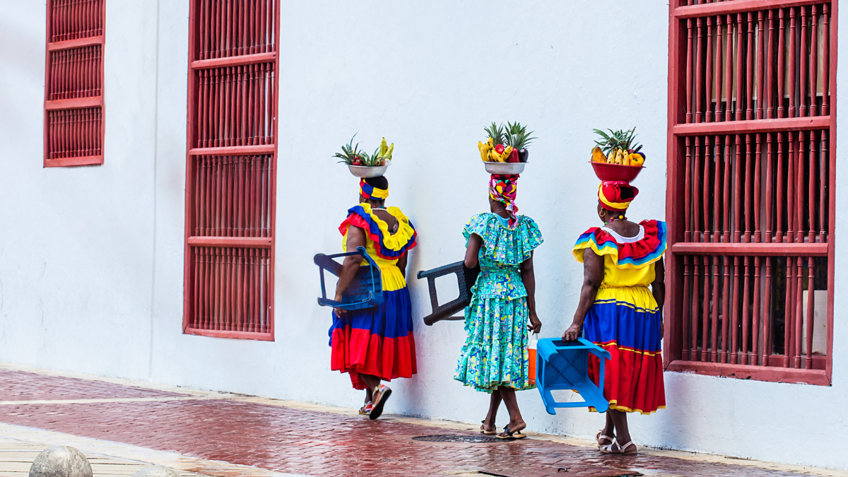 Three women in colorful traditional dresses walk along a brick path, each carrying a basket of fruit on their heads. They are passing by a white wall with red window grilles. Two women hold blue plastic chairs.