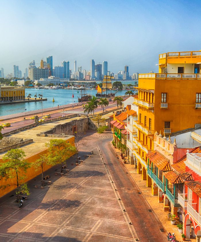 A vibrant urban scene featuring colorful colonial buildings lining a quiet street, with a waterfront view showcasing modern skyscrapers and a clear blue sky in the background.