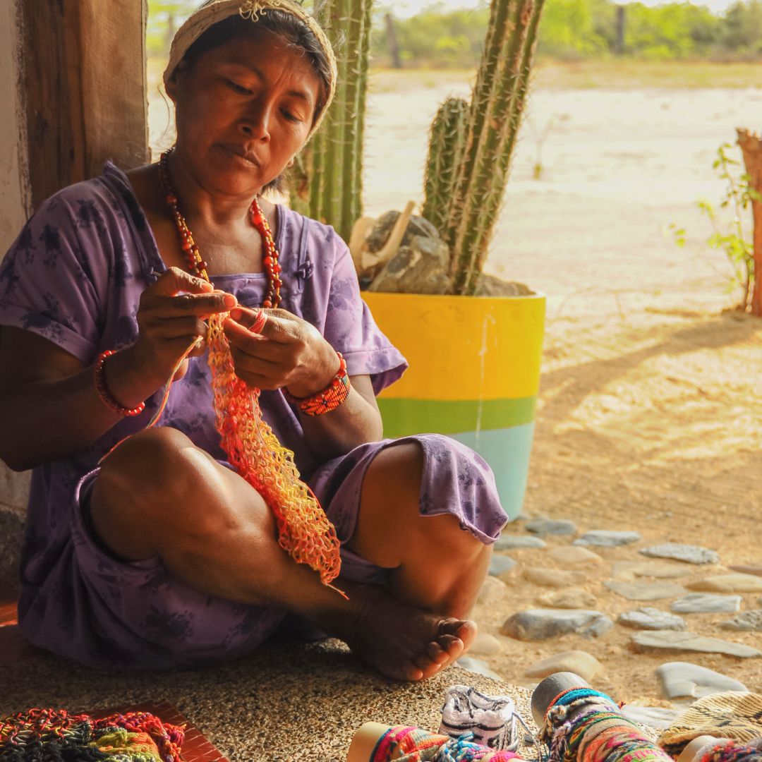 A woman sits cross-legged on the floor, focused on knitting with colorful yarn. She wears a purple dress and has traditional jewelry. In the background, there are cacti and a painted pot, suggesting a warm, sunny environment.