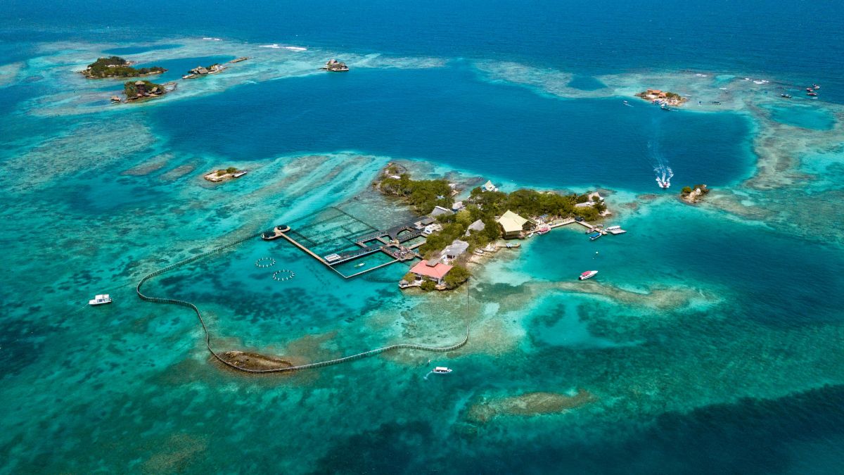 Aerial view of a tropical island surrounded by turquoise waters, featuring small rocky islets, coral reefs, and boats. The island has a cluster of buildings and structures, with some areas marked off for marine activities.