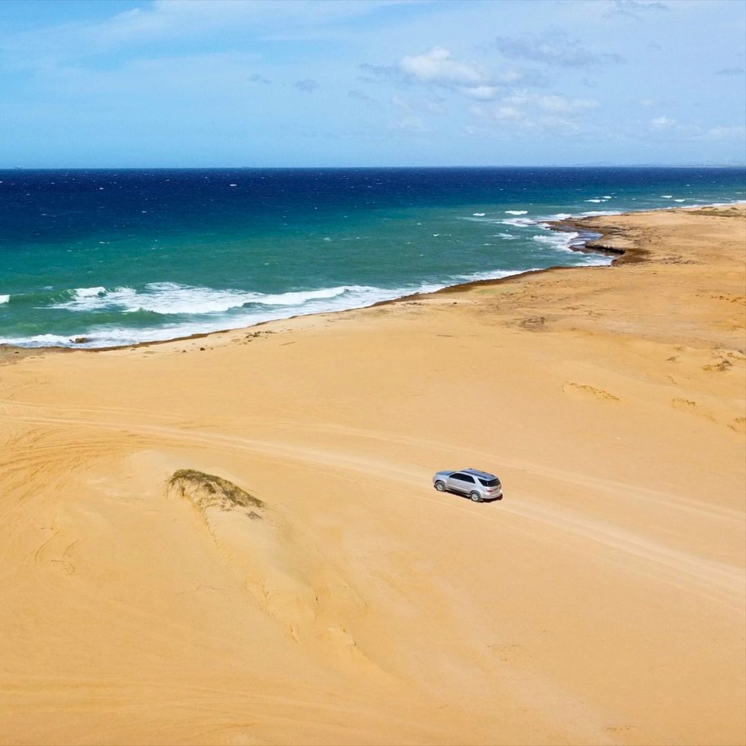 A silver car driving on a sandy path along a coastline, with vibrant blue sea and cloudy skies in the background.