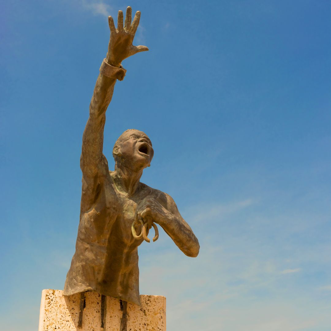 A bronze statue of a man reaching upward with one hand raised and a look of desperation on his face, set against a bright blue sky. The figure appears to be wearing a shirt and holds something in the other hand, symbolizing a plea or call for help.