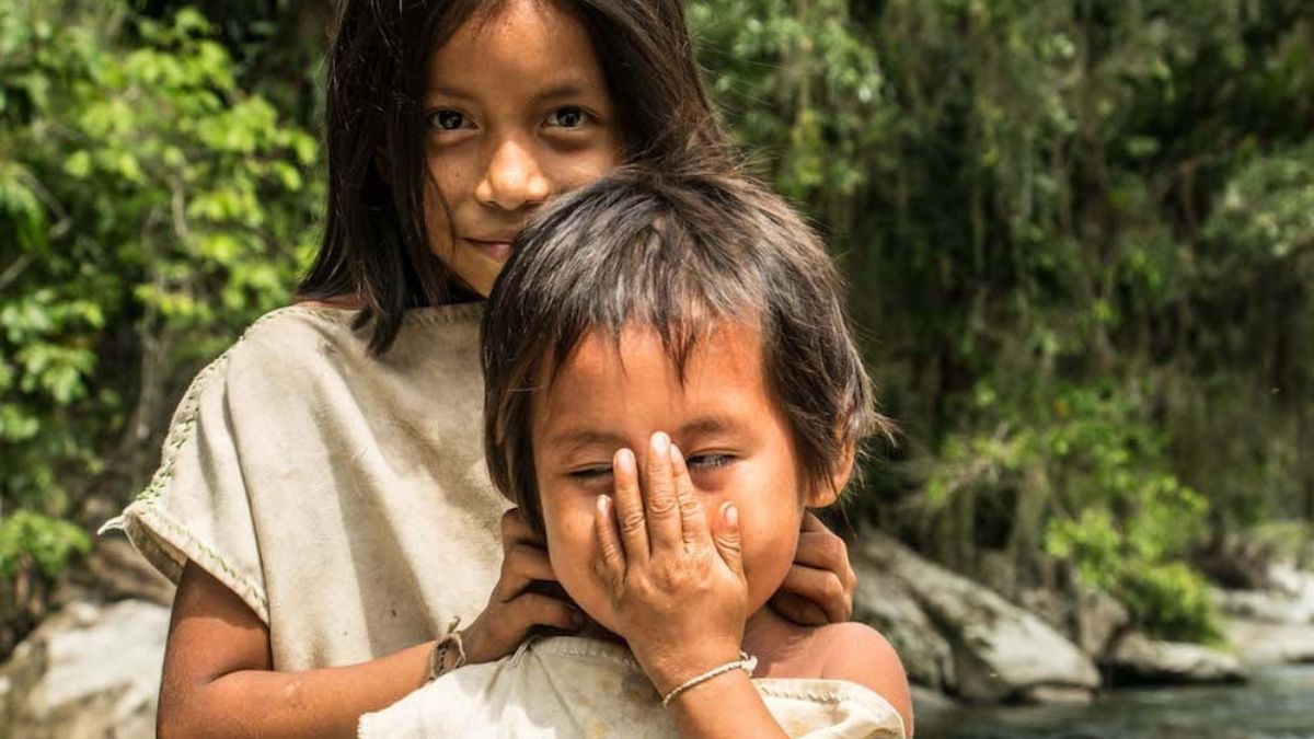 Two children play outdoors in a lush, green environment. One child stands behind the other, smiling, while the other covers their face with one hand. Both are wearing simple, natural-colored clothing.