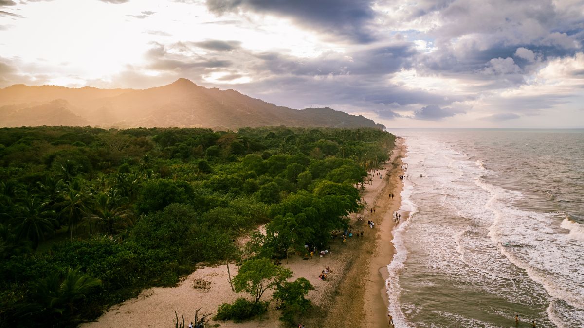 A scenic view of a coastline with lush greenery, sandy beach, and waves gently lapping at the shore. In the background, mountains are silhouetted against a cloudy sky as the sun sets. People can be seen walking along the beach and enjoying the serene environment.