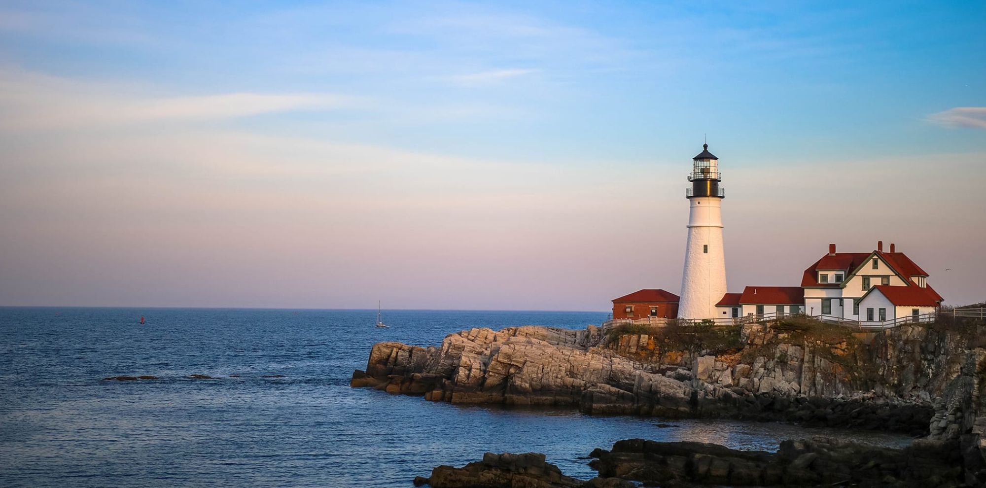 A white lighthouse with a black top stands on rocky shorelines, beside a charming red-roofed building. The sea stretches out in the background, reflecting a pastel sunset, with a sailboat visible in the distance.