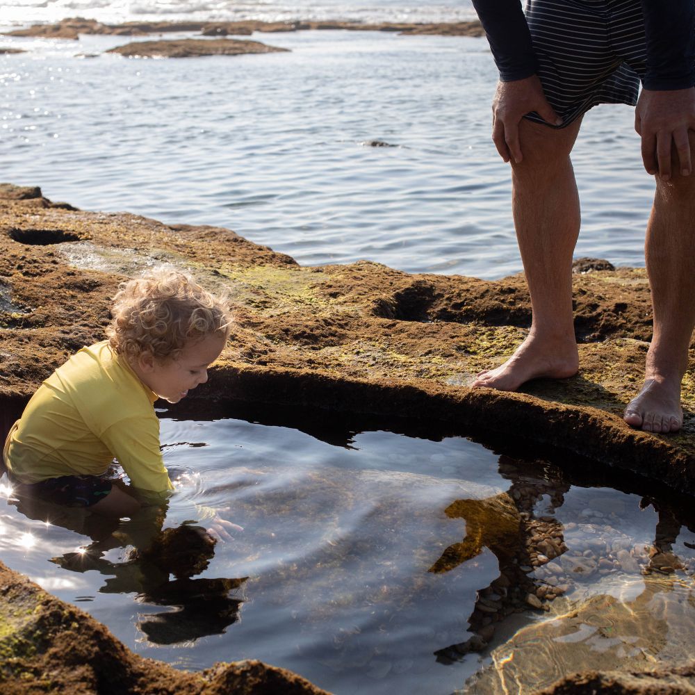 A child with curly hair is playing in a tide pool on a rocky shoreline, wearing a yellow long-sleeve top. An adult stands nearby, watching, with their bare feet on the rocks. The calm sea is visible in the background.