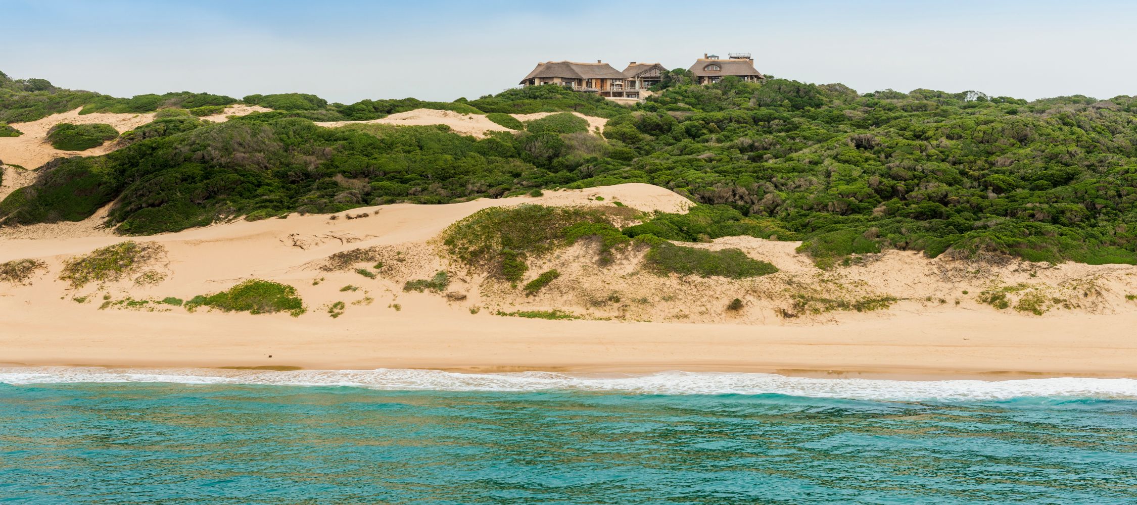 A sandy beach with gentle waves lapping at the shore. Lush greenery lines the beach, leading up to a large, multi-story house situated on a hill. The sky is clear with a soft, overcast appearance.