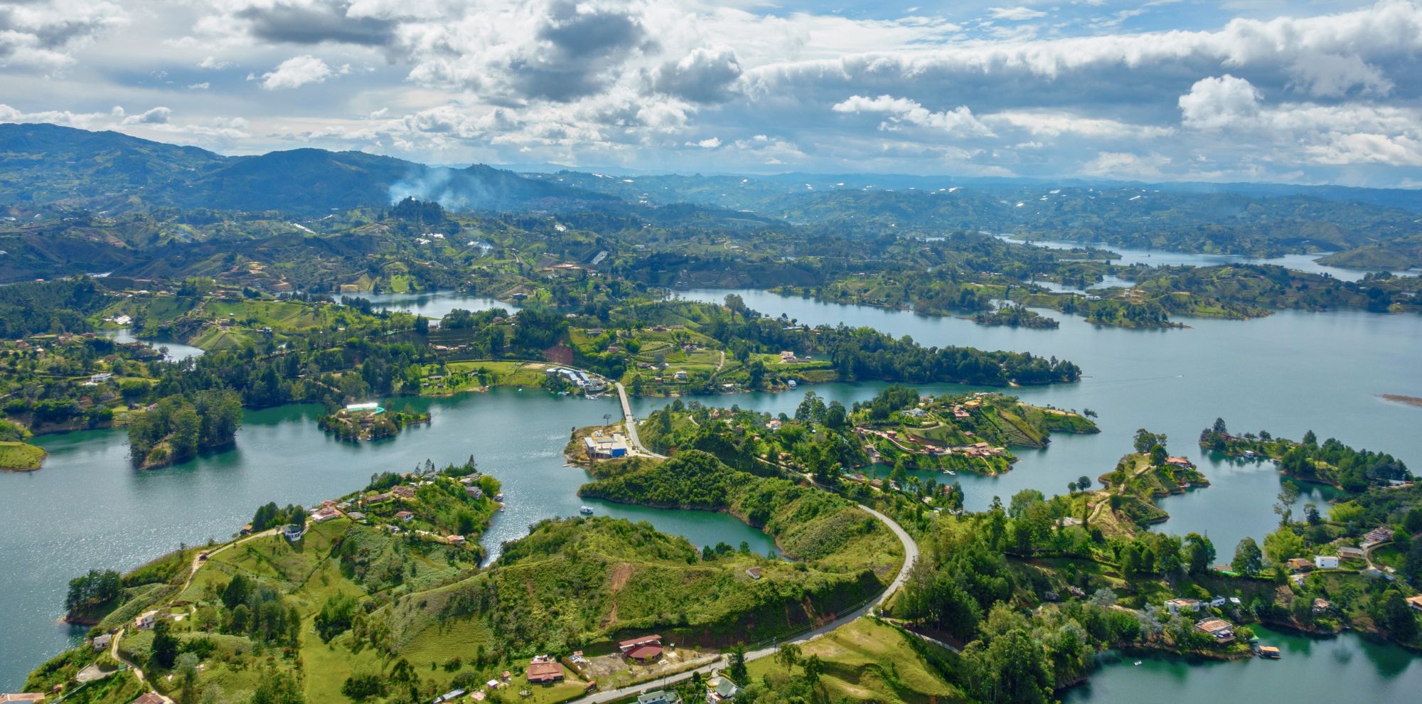 Aerial view of a lush, green landscape with a network of lakes and rivers. Rolling hills and mountains provide a scenic backdrop under a partly cloudy sky. Small homes and greenery dot the shoreline and hillsides, creating a tranquil atmosphere.