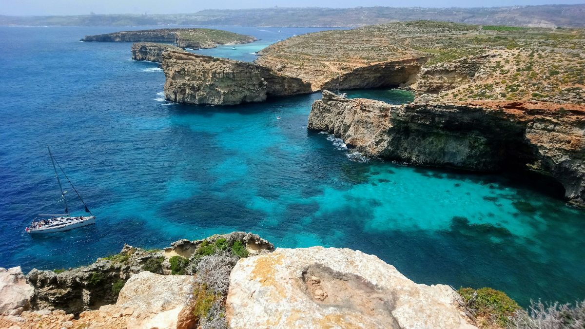 Turquoise waters surround rugged cliffs with patches of greenery. A sailboat navigates near the coastline, showcasing the natural beauty of the landscape.
