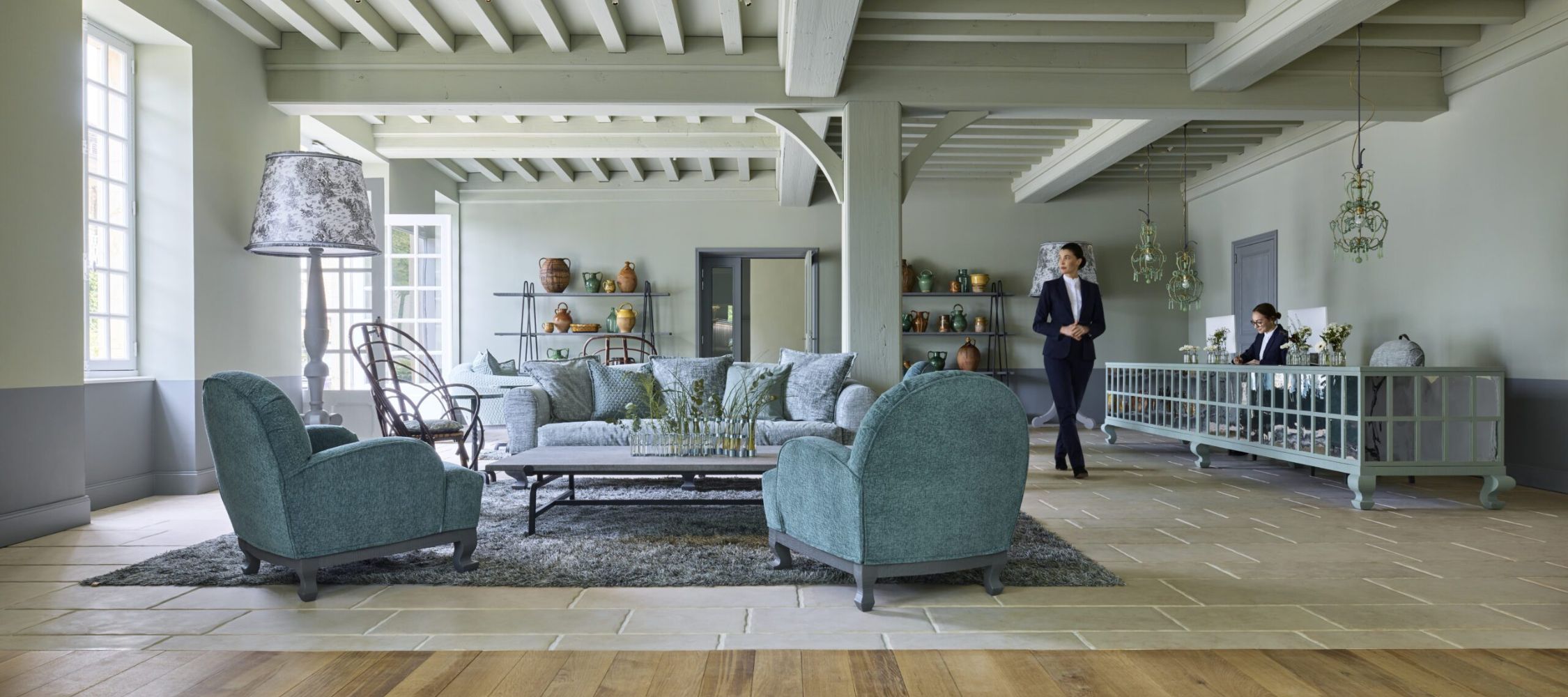 Spacious, modern living room with light green walls and wooden beams. Comfortable turquoise armchairs and a large sofa surround a coffee table on a dark rug. Decorative shelves display various items, and a person is walking while another is seated nearby.