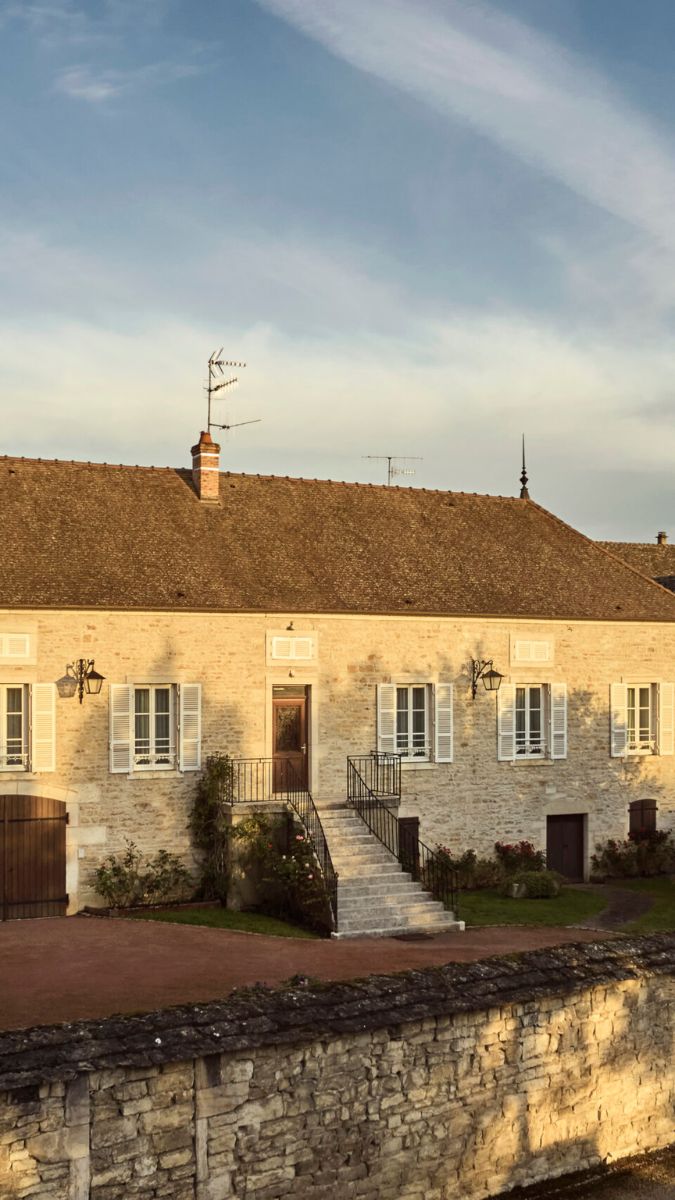 A charming stone house with a steep roof and white shutters, featuring a front staircase and garden. The surrounding area has a stone wall and a clear blue sky above.