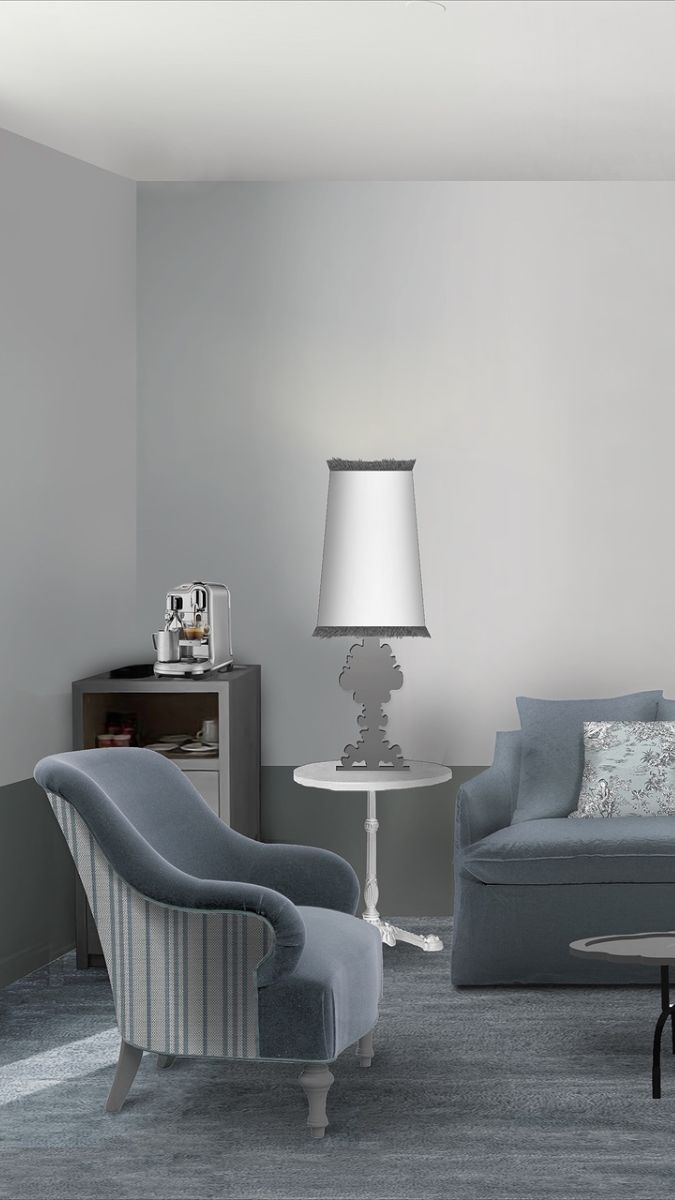 A stylish interior living space featuring a blue armchair, a sofa, a side table with an artistic lamp, and a coffee machine on a cabinet. The walls are light grey, and the floor is covered with a textured carpet.