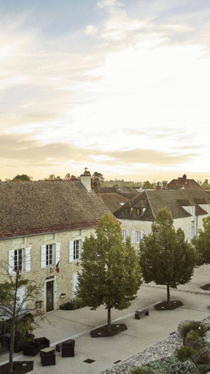 Charming village scene featuring traditional stone houses with sloped roofs under a soft sunset sky. Lush trees line a cobbled pathway, adding to the tranquil ambiance.