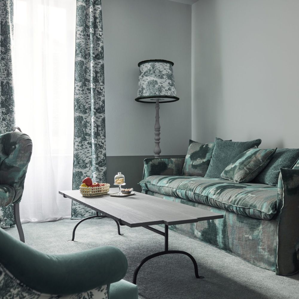 A cozy living room featuring a green and grey color scheme. There’s a plush sofa with matching cushions, a stylish coffee table, and a decorative lamp, all surrounded by patterned curtains. A small plate with strawberries sits on the table.
