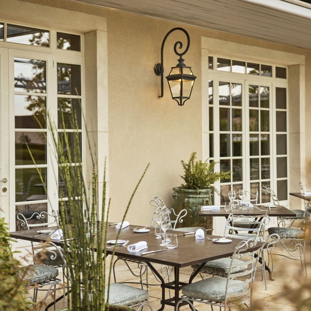 A charming outdoor restaurant setting with a stone patio, elegant metal furniture, and a vintage lantern. Potted plants and greenery enhance the inviting atmosphere.