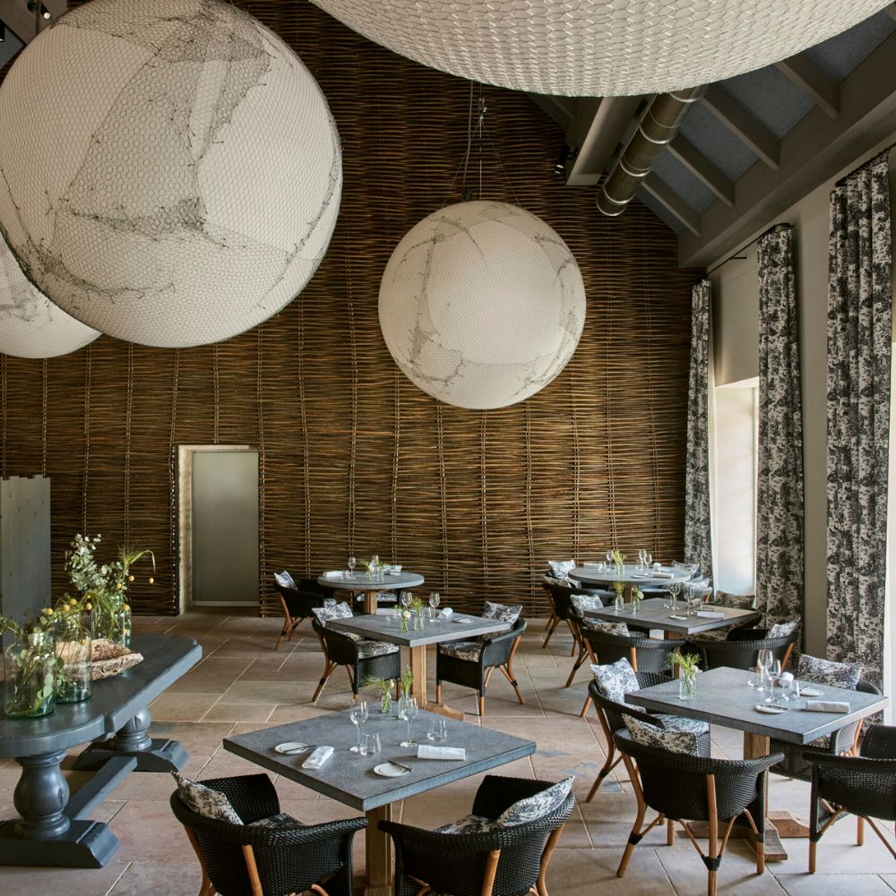 A modern restaurant interior featuring round pendant lights, rustic wooden walls, and elegantly set tables with black wicker chairs. Large windows are adorned with patterned curtains, adding warmth to the space.