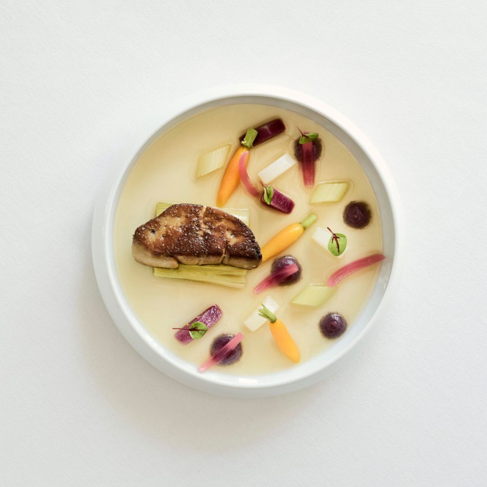 Seared foie gras served on a bed of thinly sliced vegetables, including vibrant carrots and purple radishes, in a clear broth. Garnished with fresh herbs.