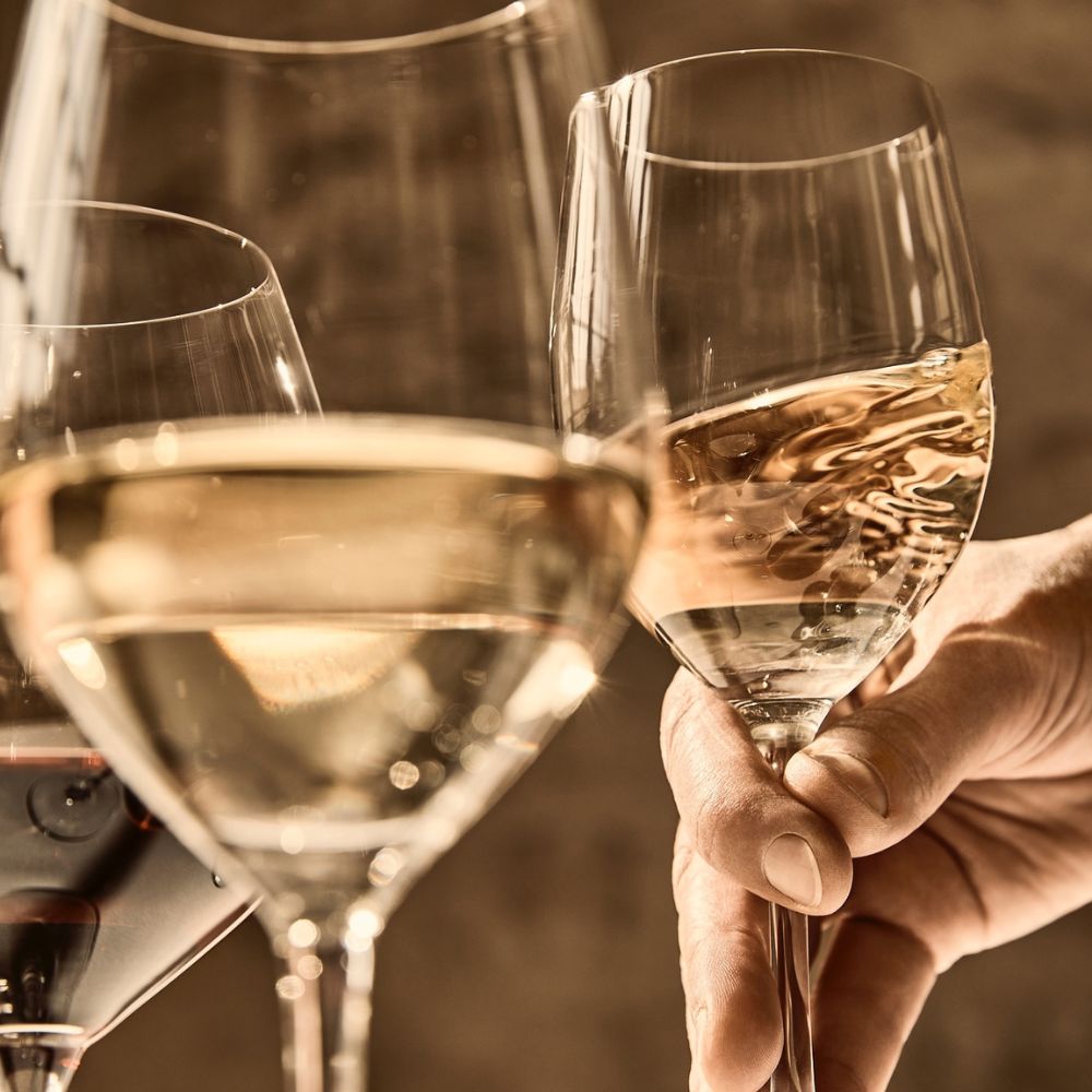 A hand holding a glass of white wine, alongside two other glasses that contain white and red wine. The background is softly blurred, emphasizing the glasses.