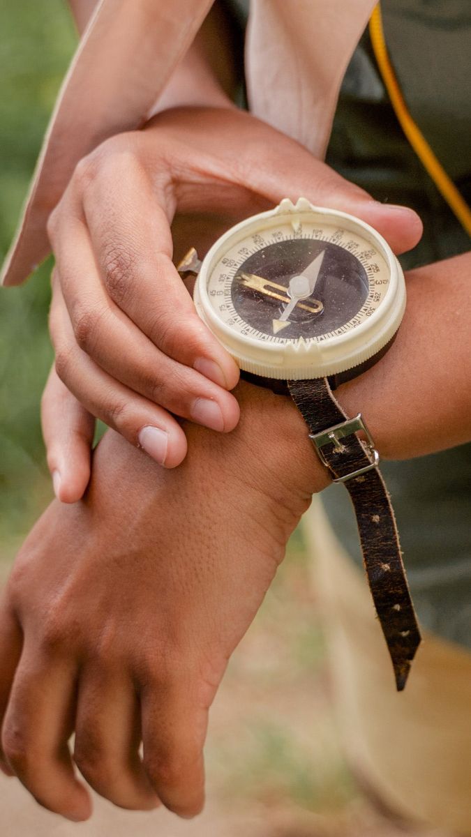 A close-up of a person's hand holding a vintage compass on their wrist. The compass features a round face with a black and gold design, and the hand is positioned in a natural outdoor setting.