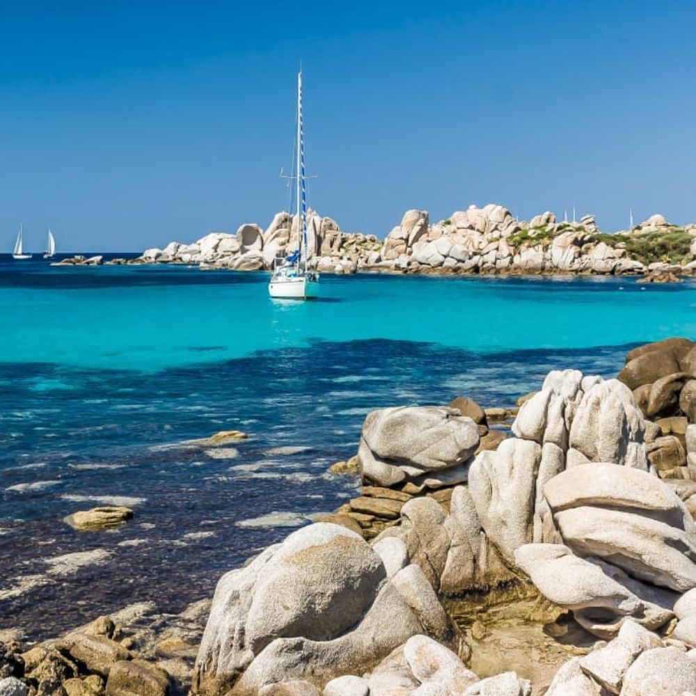 A tranquil seaside scene with clear turquoise water and a sailboat near rocky shores. Large boulders are visible in the foreground, and small sailboats can be seen in the distance under a clear blue sky.