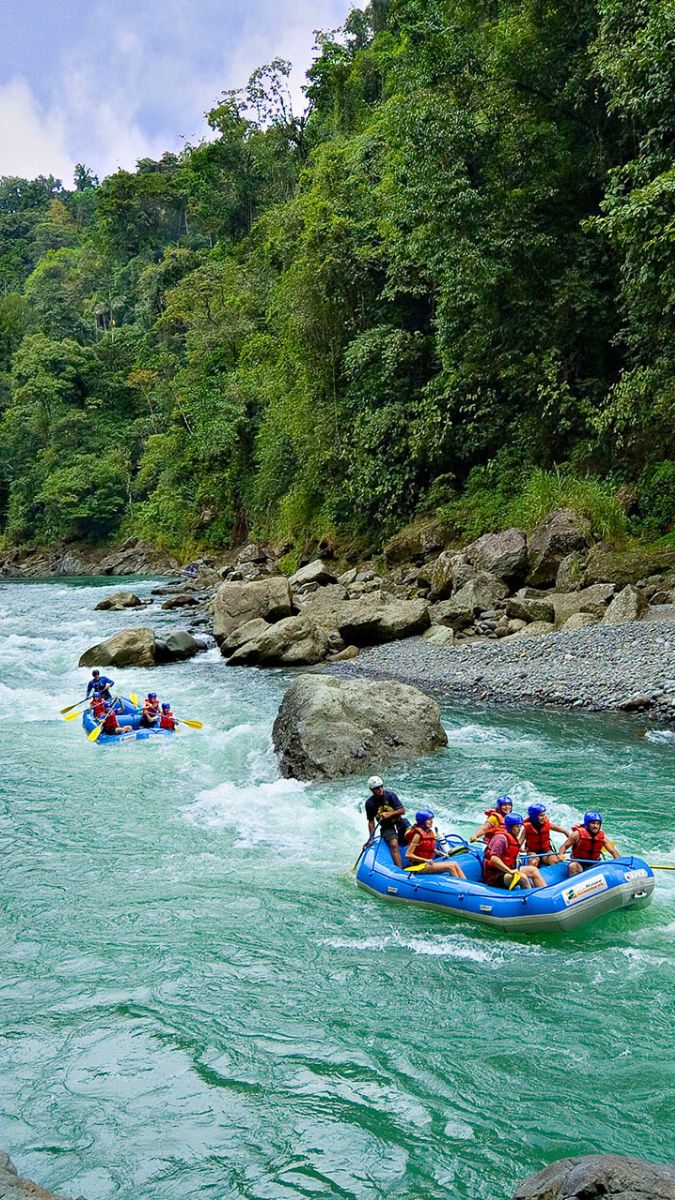 Rafts navigating a turquoise river surrounded by lush green forests and rocks. Participants are wearing life jackets and helmets as they enjoy whitewater rafting.