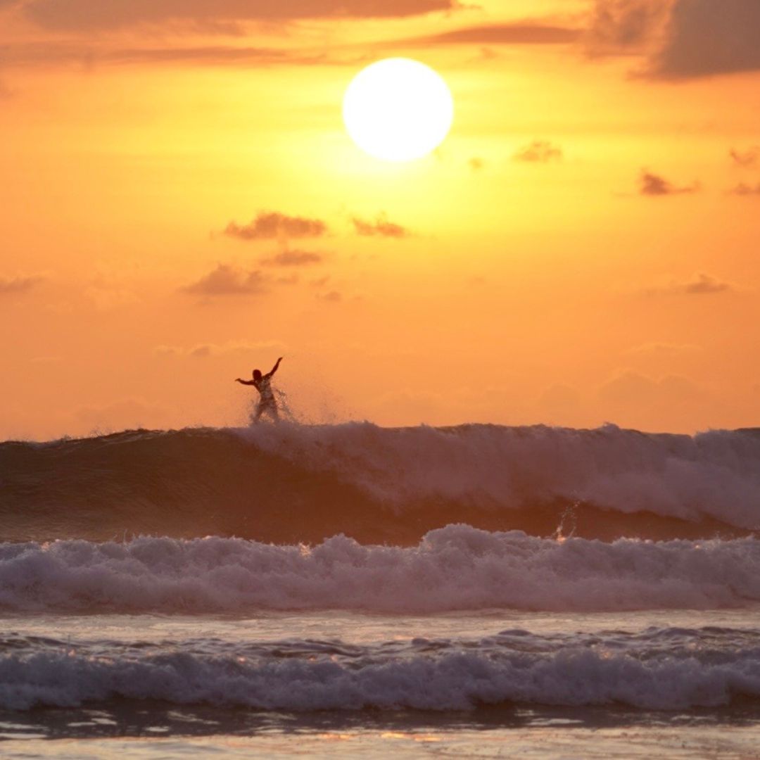 A surfer rides a wave at sunset, with a vibrant orange sky and the sun setting on the horizon.