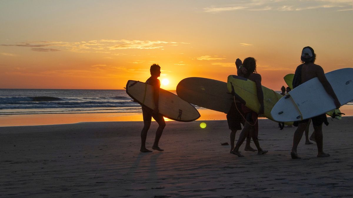 Surfers walk along the beach at sunset, carrying their surfboards while the sun sets over the ocean. The sky displays warm orange and yellow hues, reflecting on the water.