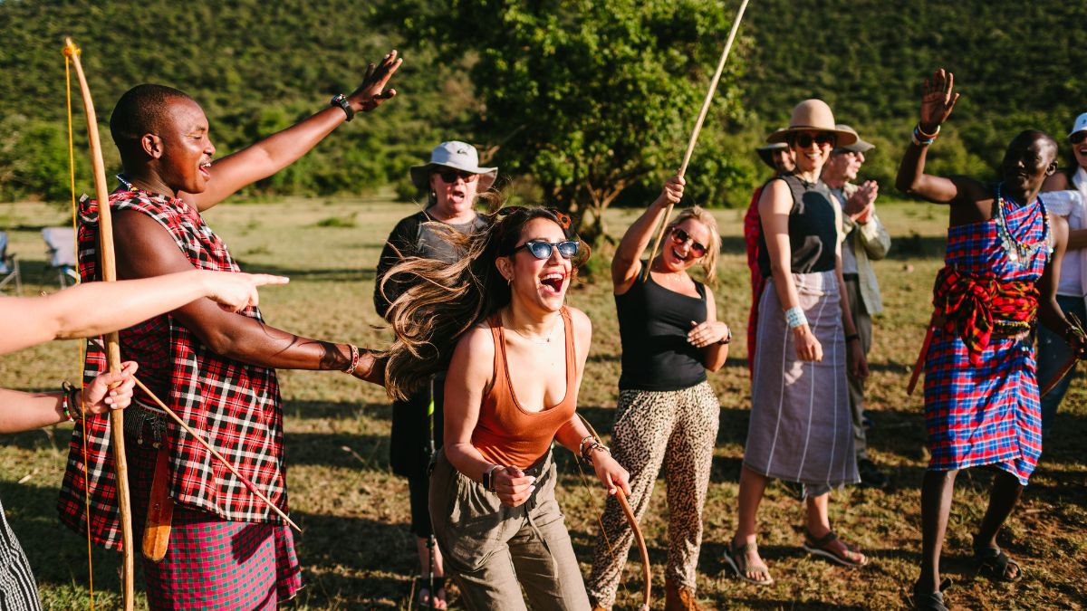 A lively group of people celebrating outdoors, with some wearing traditional attire. They appear to be dancing and laughing, enjoying the moment in a scenic landscape with green hills in the background.